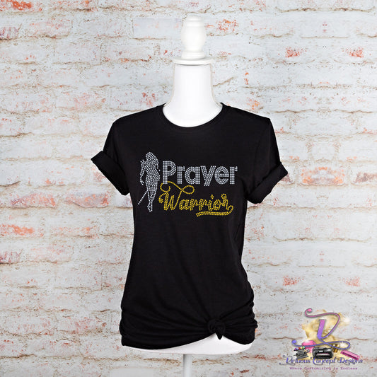 The Prayer Warrior shirt by Virtuous Concept Boutique features a black slim fit design with the words "Prayer Warrior" in white and yellow text, accented by a white silhouette. Displayed on a mannequin against a white brick wall, this bling shirt brings a sparkling touch to your faith-filled wardrobe.