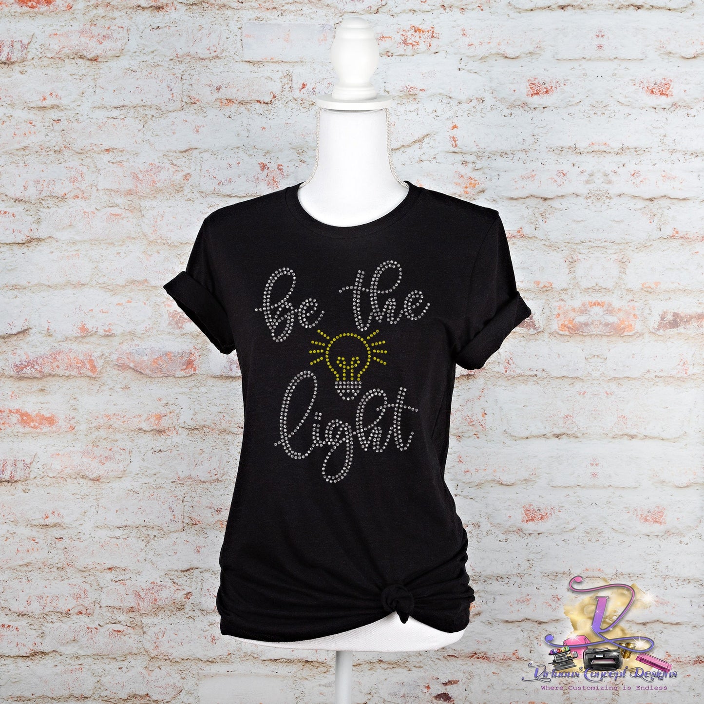 Displayed on a mannequin is the "Be the Light" slim fit t-shirt by Virtuous Concept Boutique. This black shirt features decorative text with a light bulb design and is styled with a knotted bottom, adding flair against a white brick wall backdrop.