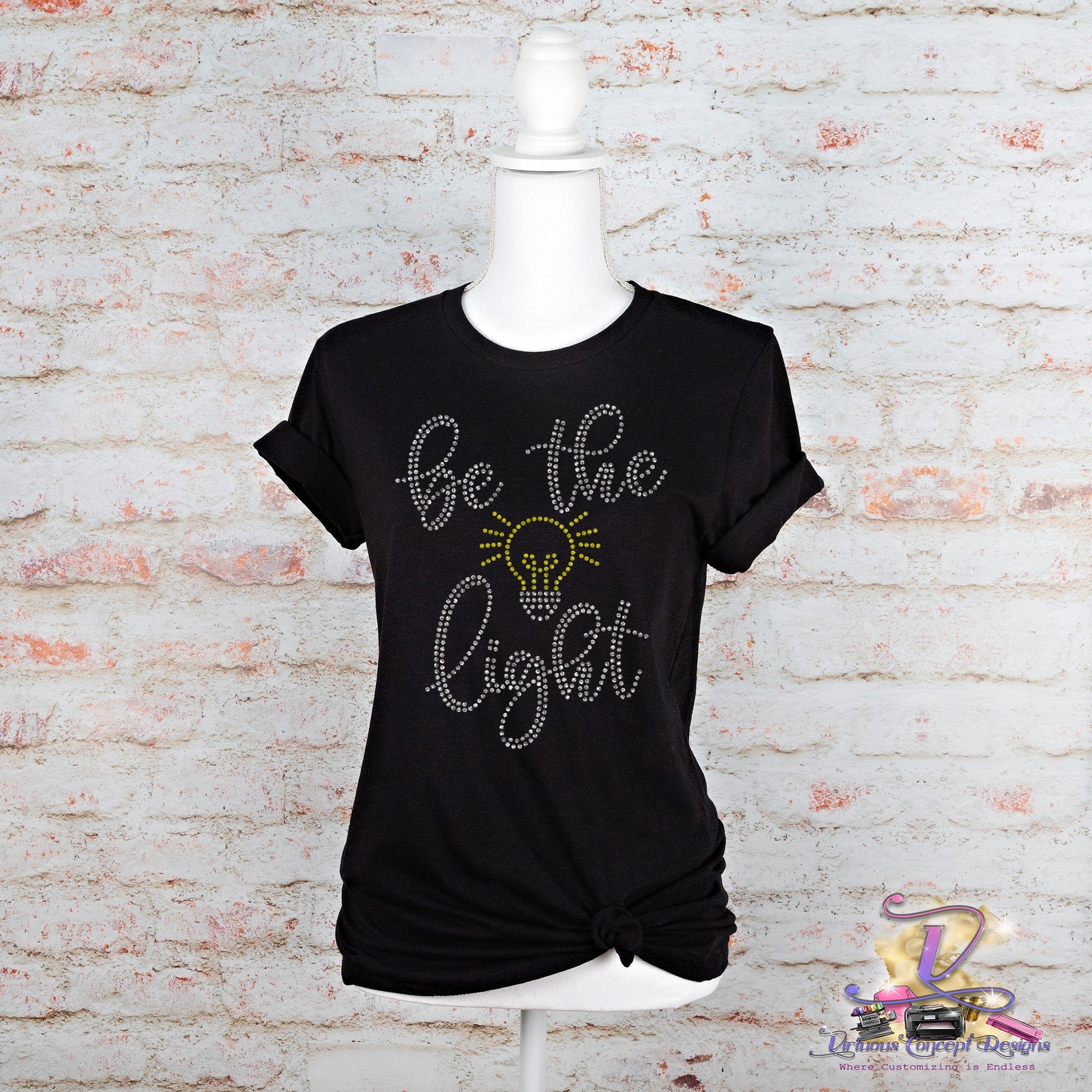 Displayed on a mannequin is the "Be the Light" slim fit t-shirt by Virtuous Concept Boutique. This black shirt features decorative text with a light bulb design and is styled with a knotted bottom, adding flair against a white brick wall backdrop.
