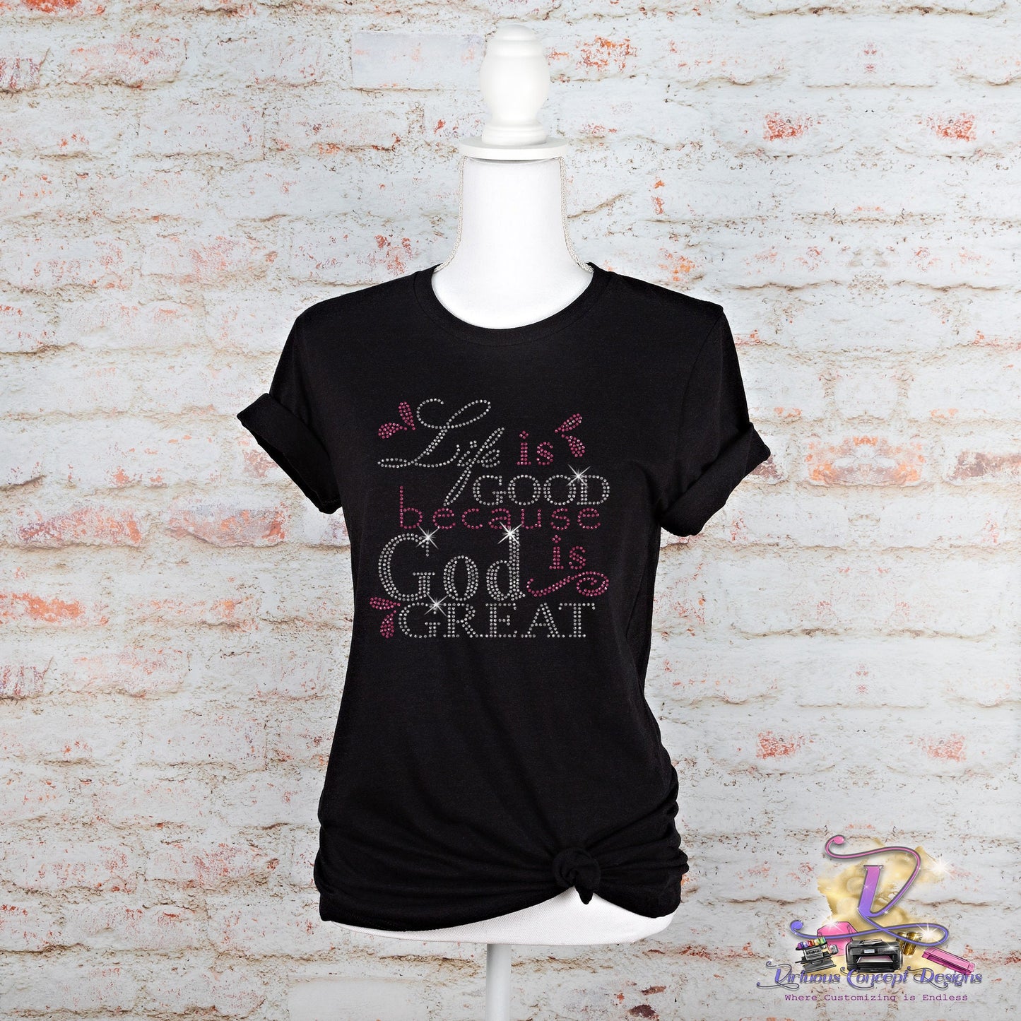 A black slim fit T-shirt from Virtuous Concept Boutique, titled "Life is good," is displayed on a mannequin against a white brick background. The shirt features the phrase "Life is good because God is great" in decorative fonts, embellished with small pink and white heart designs and has rolled sleeves.