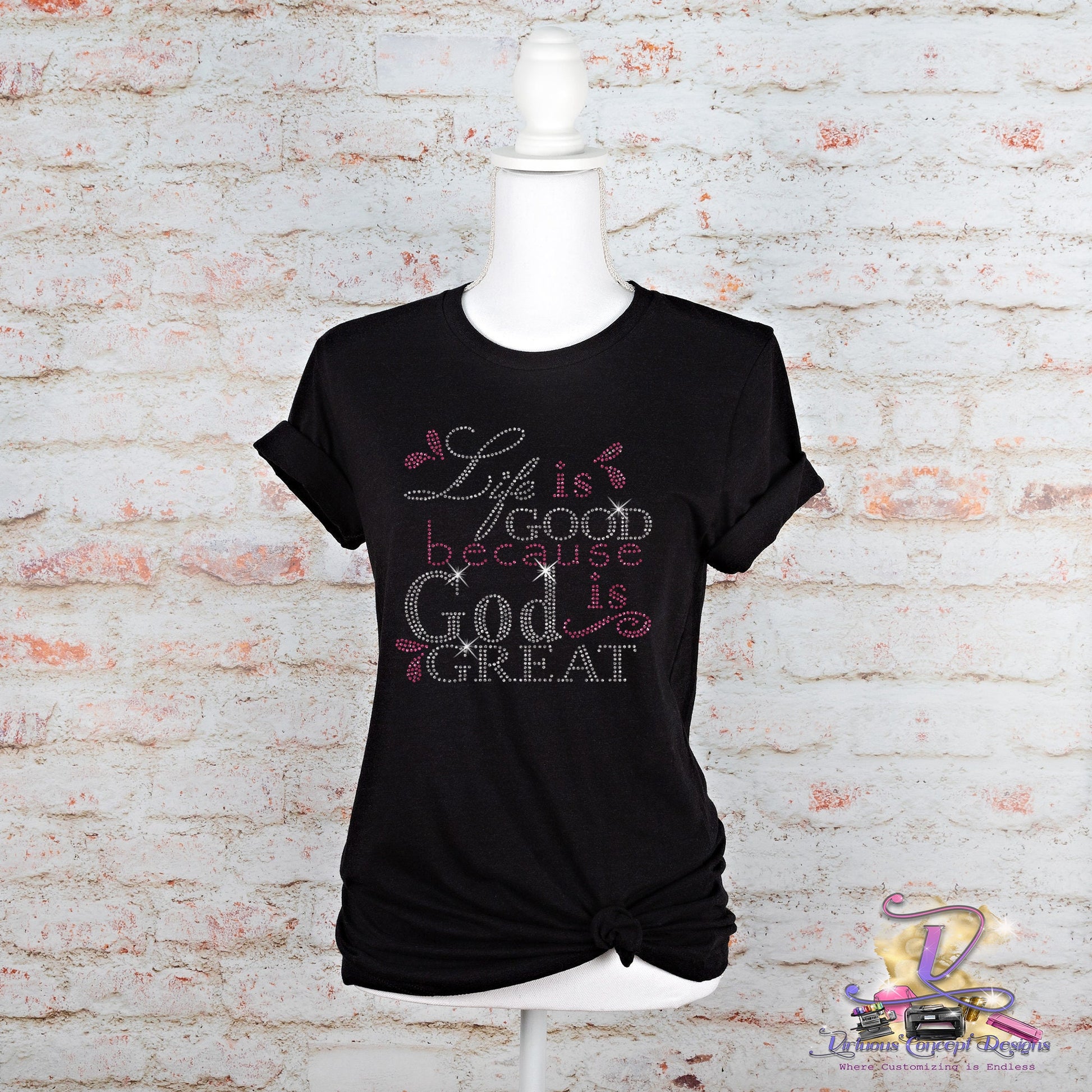 A black slim fit T-shirt from Virtuous Concept Boutique, titled "Life is good," is displayed on a mannequin against a white brick background. The shirt features the phrase "Life is good because God is great" in decorative fonts, embellished with small pink and white heart designs and has rolled sleeves.