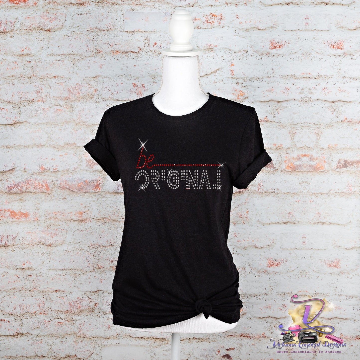 A mannequin showcases the Be Original t-shirt from Virtuous Concept Boutique, featuring dazzling rhinestone lettering. With its stylish knot at the bottom, this trendy piece stands out against a white brick wall, inviting you to sparkle with confidence.