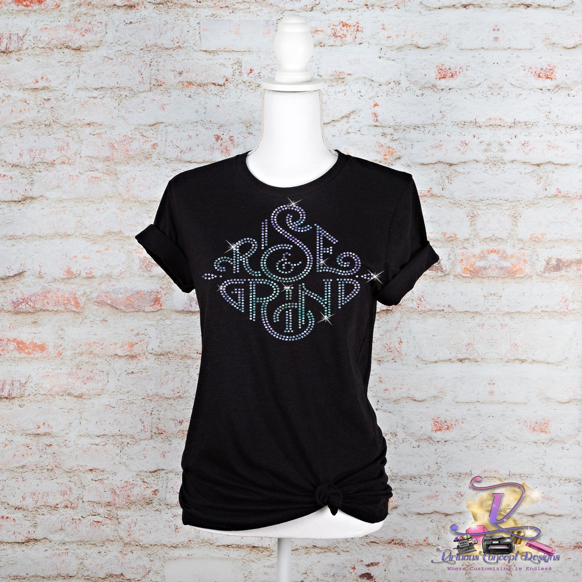 Displayed on a white mannequin against a textured white brick background is the "Rise & Grind" shirt by Virtuous Concept Boutique. This sleek black top features rolled sleeves and a knotted hem, with an eye-catching swirling design crafted from colorful dots.