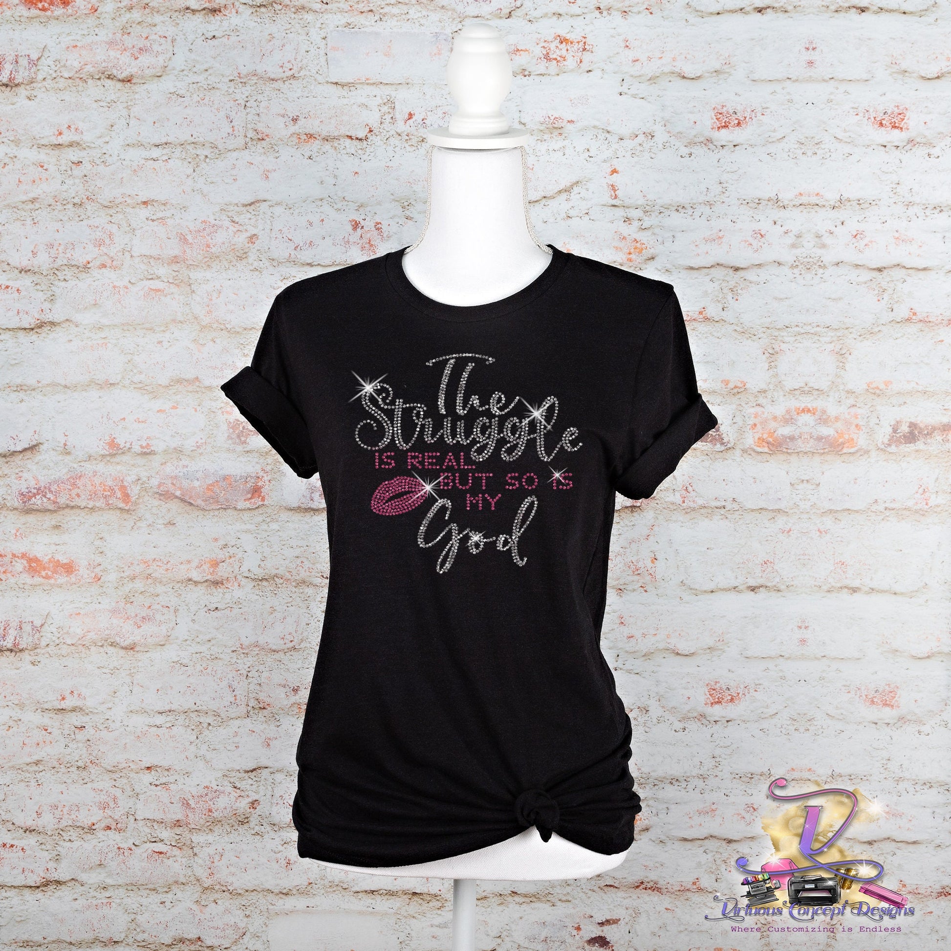 Against a white brick wall backdrop, a mannequin displays "The Struggle is Real" shirt by Virtuous Concept Boutique. This black T-shirt features glittery script that reads, "The struggle is real, but so is my God," and has a chic knotted side detail that catches the eye.